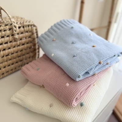 Suéteres de malha azul claro, rosa e branco com pompons coloridos dobrados junto a bolsa de palha trançada