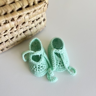 Sapatinhos de bebé em crochet azul claro com tiras, junto a cesta de palha trançada