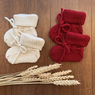 Botinhas de bebé em malha bege e vermelha com laços, sobre madeira com espigas de trigo