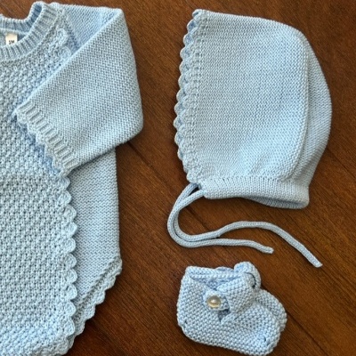 Conjunto de roupa de bebé azul claro em fio com camisola, touca e peúga sobre madeira