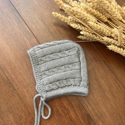 Touca cinzenta de malha com padrão trançado e cordão, sobre madeira com ramo de trigo ao lado