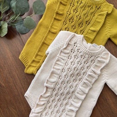 Peças de roupa de bebé amarelo mostarda e creme em malha rendada com folhos, sobre madeira com folhas verdes