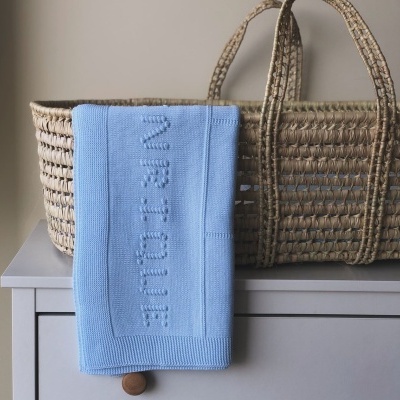 Manta azul com texto 'MR. TOQUE' pendurada numa cómoda cinzenta com cesto de palha atrás