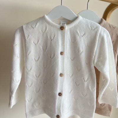 Casaco de malha branco para bebé com botões de madeira pendurado num cabide.