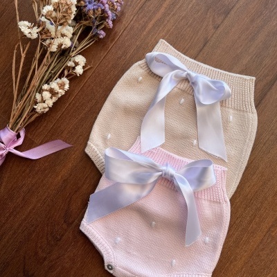 Dois bodies de bebé em malha, bege e rosa, com laços na cintura, e ramo de flores secas ao lado