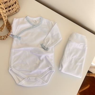 Roupa de bebé branca e azul claro com body e calças em cima de uma mesa branca