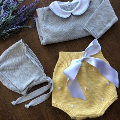 Conjunto de bebé com camisola cinza, calção amarelo com laço branco e gorro cinza sobre madeira