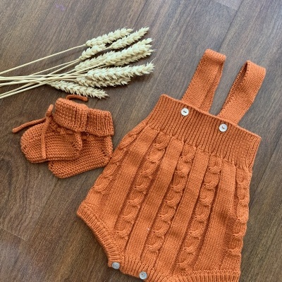 Macacão e botas de bebé tricotados em laranja sobre chão de madeira com espigas de cereal