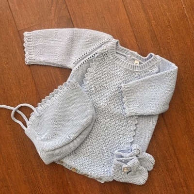 Conjunto de bebé em malha azul clara com camisola, gorro e pantufas sobre chão de madeira.