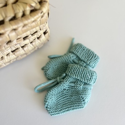 Par de luvas para bebé azuis em malha, com textura de tricô, ao lado de uma cesta de palha