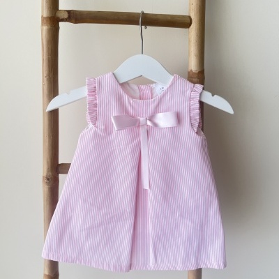 Vestido de bebé rosa com riscas brancas e laço de cetim pendurado numa estrutura de madeira.