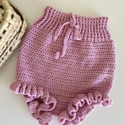 Calções cor-de-rosa de crochet com cordão e folhos, sobre uma mesa branca junto a uma cesta de verga