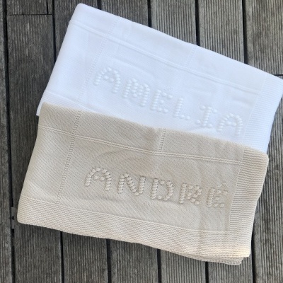 Duas toalhas de banho, uma branca com nome AMELIA e outra bege com nome ANDRE, sobre pavimento de madeira.