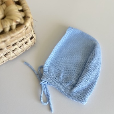 Touca azul clara de malha fina com fitas, sobre mesa branca, ao lado de cesto de palha