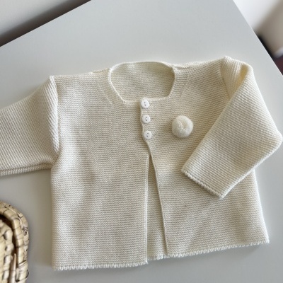 Casaco creme de malha para bebé com botões e pompom numa mesa branca