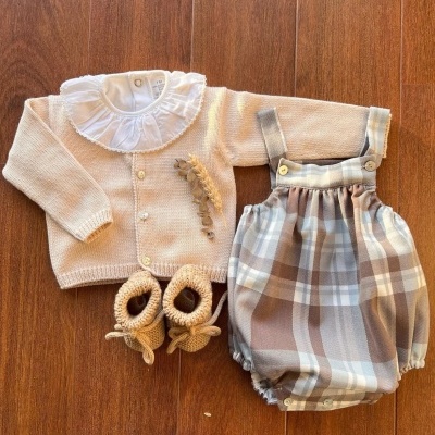 Conjunto de roupa de bebé com casaco bege, body branco com gola e patas xadrez bege e castanho sobre madeira.