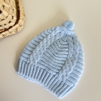 Gorro azul claro de malha com pompom colocado em superfície branca ao lado de cesto trançado