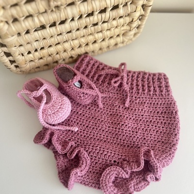 Conjunto de roupa de bebé em croché cor-de-rosa com calça e sapatinhos ao lado de uma cesta de verga.