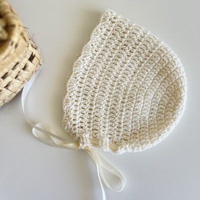Touca infantil de croché branco com fita branca ao lado de cesta de palha.