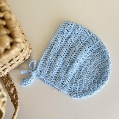 Gorro de crochet azul claro com cordão em superfície branca ao lado de cesto de fibras naturais