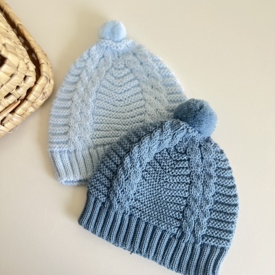 Duas gorros de malha azuis com pompom sobre mesa branca, com cesta de palha ao lado.