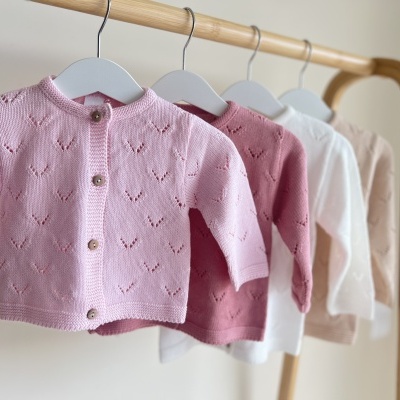 Três casacos de malha para bebé de cores rosa claro, rosa escuro e branco pendurados em cabides de madeira