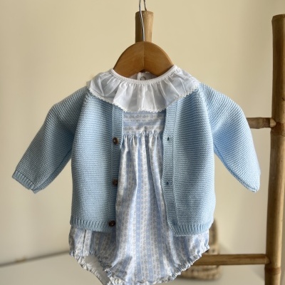 Roupa de bebé azul claro com casaquinho de malha e macacão floral branco e azul pendurado em cabide de madeira
