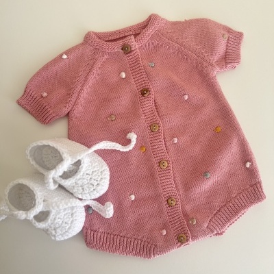 Macacão de bebé rosa com botões e detalhes coloridos ao lado de sapatinhos brancos de croché.