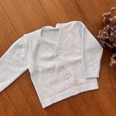 Casaco de malha branco para criança com botões, colocado sobre chão de madeira com flores secas ao lado