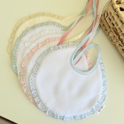Babetes brancos com bordas em renda colorida e fitas para amarrar, dispostos em leque ao lado de um cesto trançado