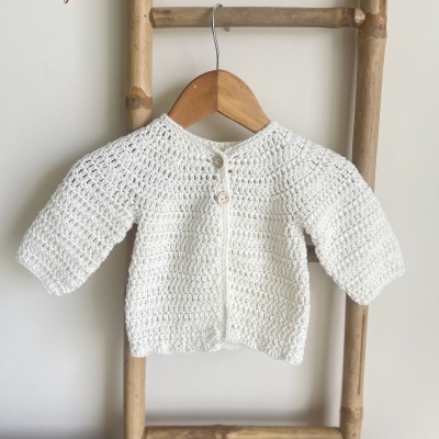 Casaco bebé de malha crochê branco pendurado em cabide na escada de bambu