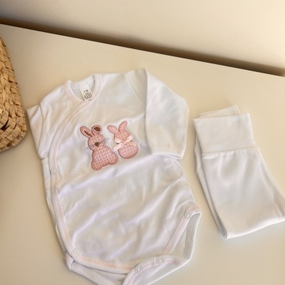 Body bebé branco com coelhos cor-de-rosa bordados e peça branca ao lado sobre superfície clara