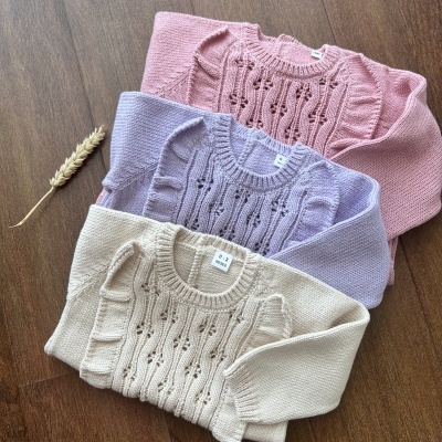 Três camisolas de malha nas cores rosa, lilás e bege sobre chão de madeira escura.