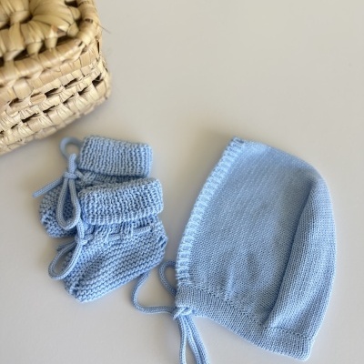 Gorro e botinhas de bebé em malha azul claro sobre mesa branca com cesto de palha ao lado