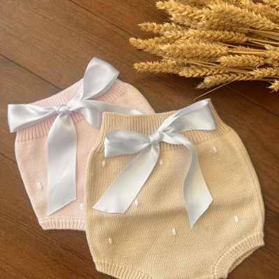Dois calções de bebé em malha rosa e bege com laços brancos, sobre madeira com espigas secas