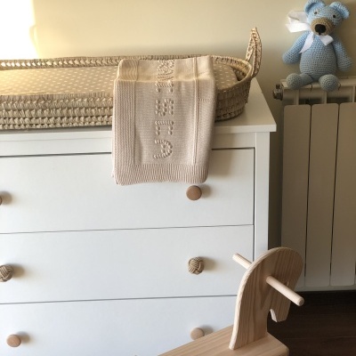 Cómoda branca com cesto de vime e cobertor bege, cadeira de balanço de madeira em forma de cavalo e urso azul de croché