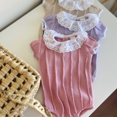 Três bodies de bebé em malha com gola de renda branca, em rosa, lilás e bege, sobre uma mesa branca com uma cesta de palha próxima.