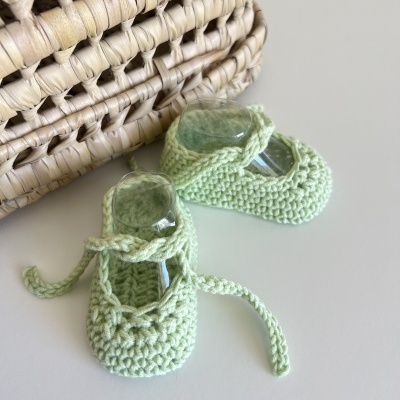 Sapatinhos de bebé em malha verde clara com fitas e bolsa de palha ao lado
