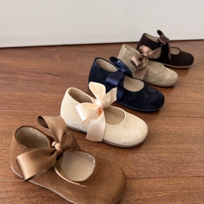 Sapatos de criança em camurça com laços de cetim, várias cores, sobre chão de madeira.