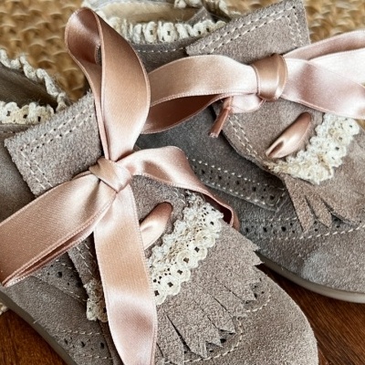 Sapatos infantis bege com fita rosa e renda branca sobre tapete de juta