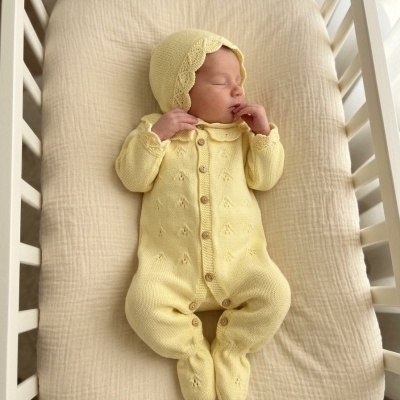 Bebé a dormir com macacão de malha amarelo com capuz num berço branco.