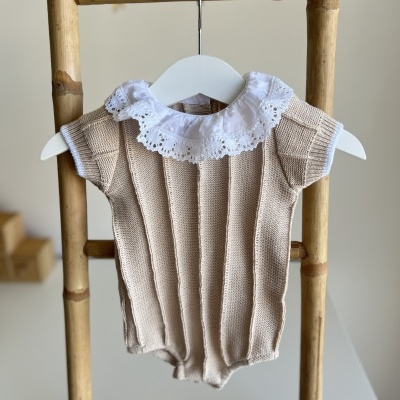 Macacão de bebé bege com gola branca em renda pendurado num cabide de plástico branco numa estrutura de bambu.