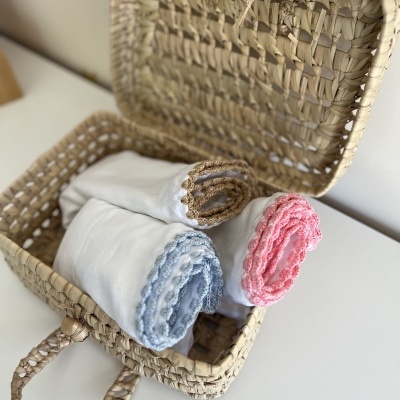 Cesta de verga aberta com três panos de tecido branco com bordas rendadas em azul, bege e rosa