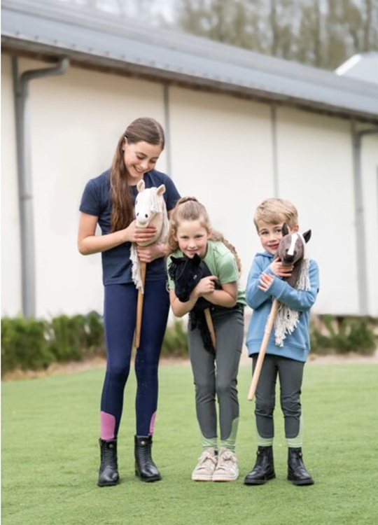 Hobby Horse HKM 'Professional Slim Line' Três crianças com brinquedos de cavalo de pau num campo de relva.