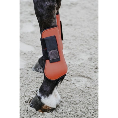 Proteção laranja para perna de cavalo com velcro preto numa perna de cavalo escura com pelo branco no casco, sobre chão de areia.