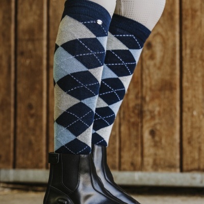 Meias justas com padrão de losangos em cinza e azul usadas com botas pretas e calças brancas, fundo de madeira