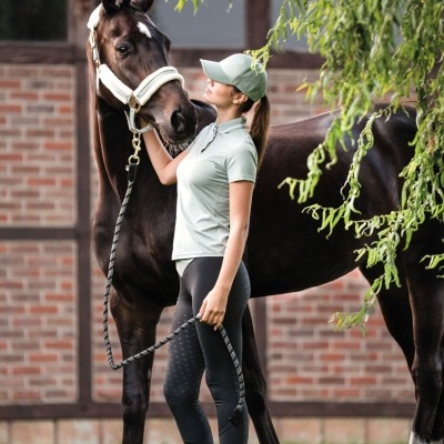 Mulher com roupa desportiva bege claro e calças pretas junto a cavalo castanho escuro perto de muro de tijolos