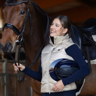 Mulher com colete bege claro, capacete azul escuro e cavalo castanho com sela preta numa área de estábulos