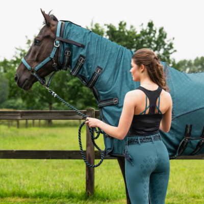Cavalo com cobertura azul esverdeada e mulher com roupa desportiva preta e azul esverdeada junto a cerca de madeira.