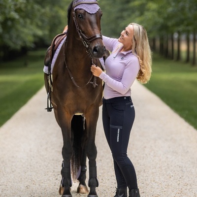 Mulher loira com roupa de equitação junto a cavalo castanho com proteção lilás nas orelhas.